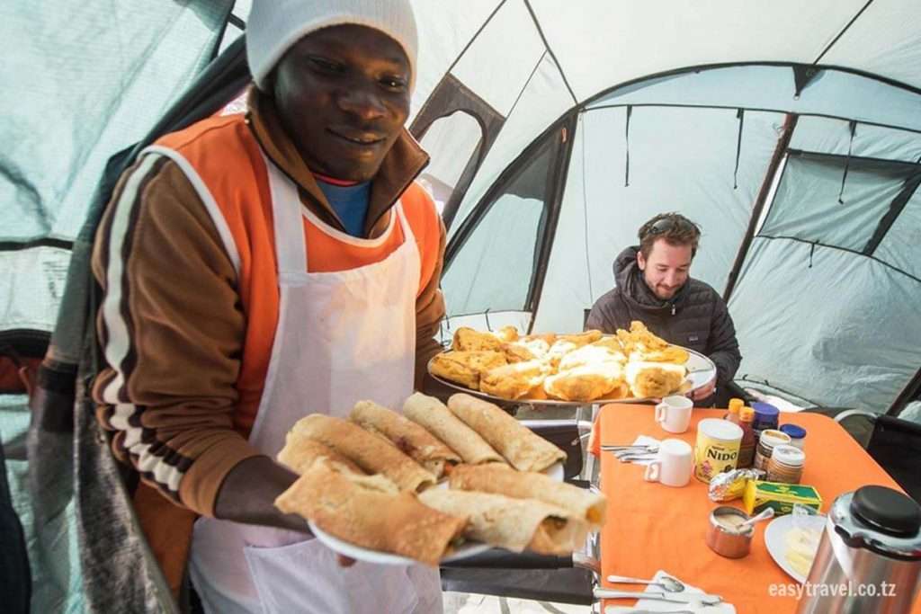 Une personne coiffée d'un bonnet et portant un tablier sert des crêpes et des beignets dans une tente qui rappelle un camp de base du Kilimandjaro. Un homme est assis à une table avec divers condiments, du thé et un appareil photo. L'atmosphère est chaleureuse et détendue. - Easy Travel Tanzania