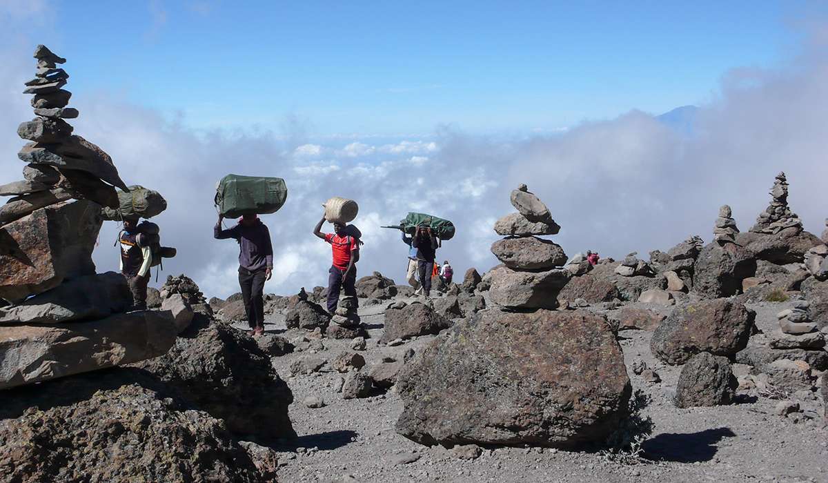 Porters carrying supplies on their heads ascend Kilimanjaros rocky path. The foreground features stacked cairns, and clouds drift below the hikers under a clear blue sky. - Easy Travel Tanzania