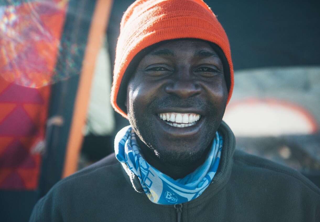 Frank Mboya, sporting an orange beanie and a blue patterned neck gaiter, smiles broadly. They are outdoors, with the background blurred into hints of colorful patterns and sunlight. - Easy Travel Tanzania