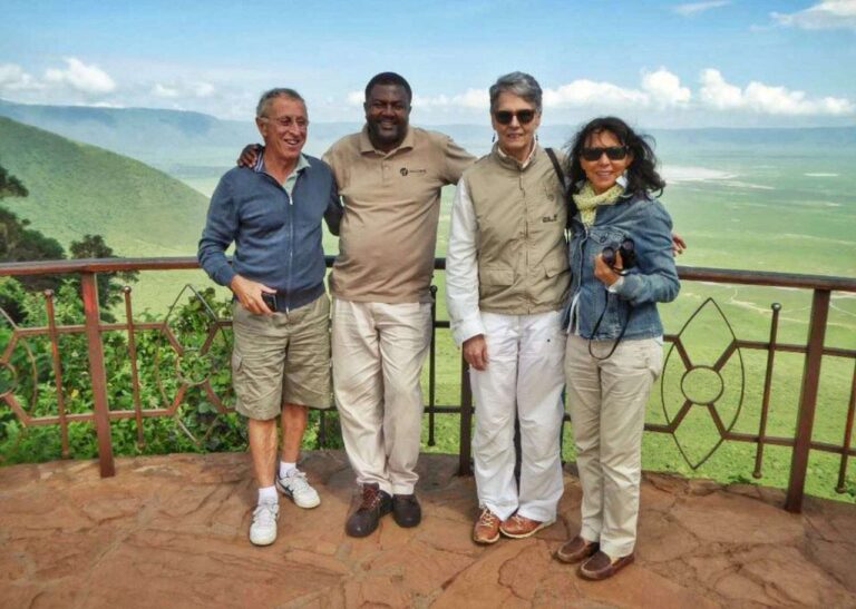 Four friends, including Gerald, stand smiling on a scenic balcony overlooking a lush green landscape and distant hills. The sky is clear with a few clouds. Casually dressed, one holds a camera as they huddle together, relishing the breathtaking view. - Easy Travel Tanzania