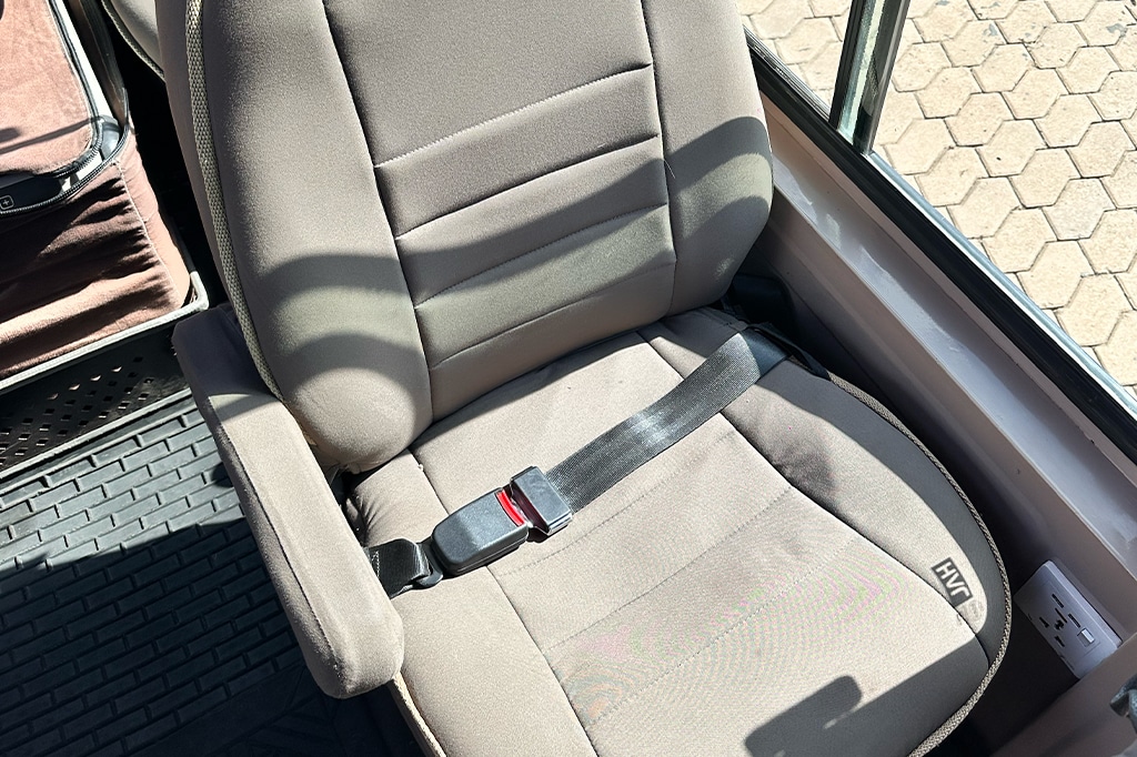 A gray bus drivers seat with armrests and a seatbelt, reminiscent of those in a safari vehicle, is placed on a rubber mat flooring. Sunlight casts shadows on the seat, which is located near a large window. - Easy Travel Tanzania
