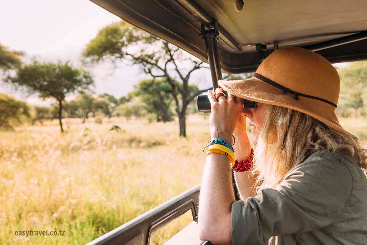 Une femme portant un chapeau et des bracelets colorés regarde à travers des jumelles tout en étant assise dans un véhicule de safari, explorant l'un des meilleurs endroits pour observer le paysage de savane herbeuse avec des arbres dispersés. - Easy Travel Tanzania