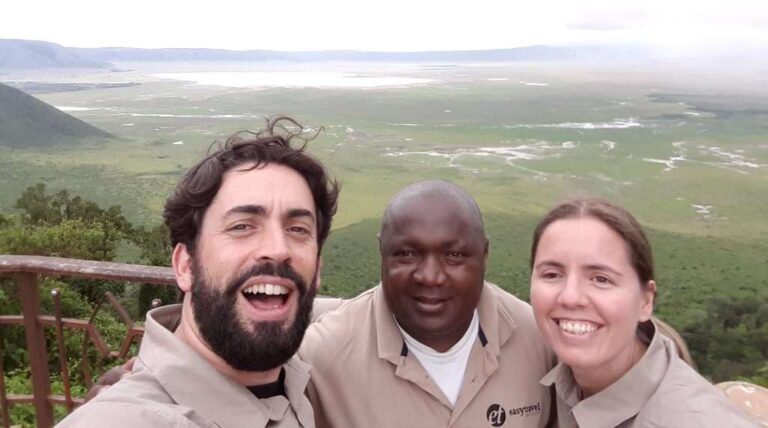 Three people, including Claus and Nikita, smiling in khaki-colored shirts, pose for a selfie against a scenic backdrop of green fields and a distant water body. Theyre standing near a wooden railing with expansive views of the landscape. - Easy Travel Tanzania