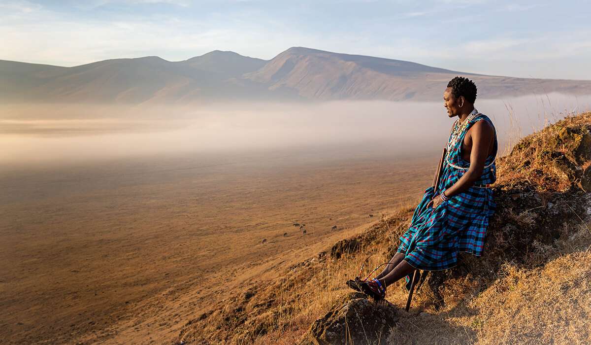 タンザニアの部族の伝統衣装をまとった人物が岩棚に座り、澄み切った空の下、広大な霧の谷と遠くの山々を見下ろしています。静かで絵のように美しいこの光景は、内省と自然の美しさの瞬間を捉えています。 - Easy Travel Tanzania