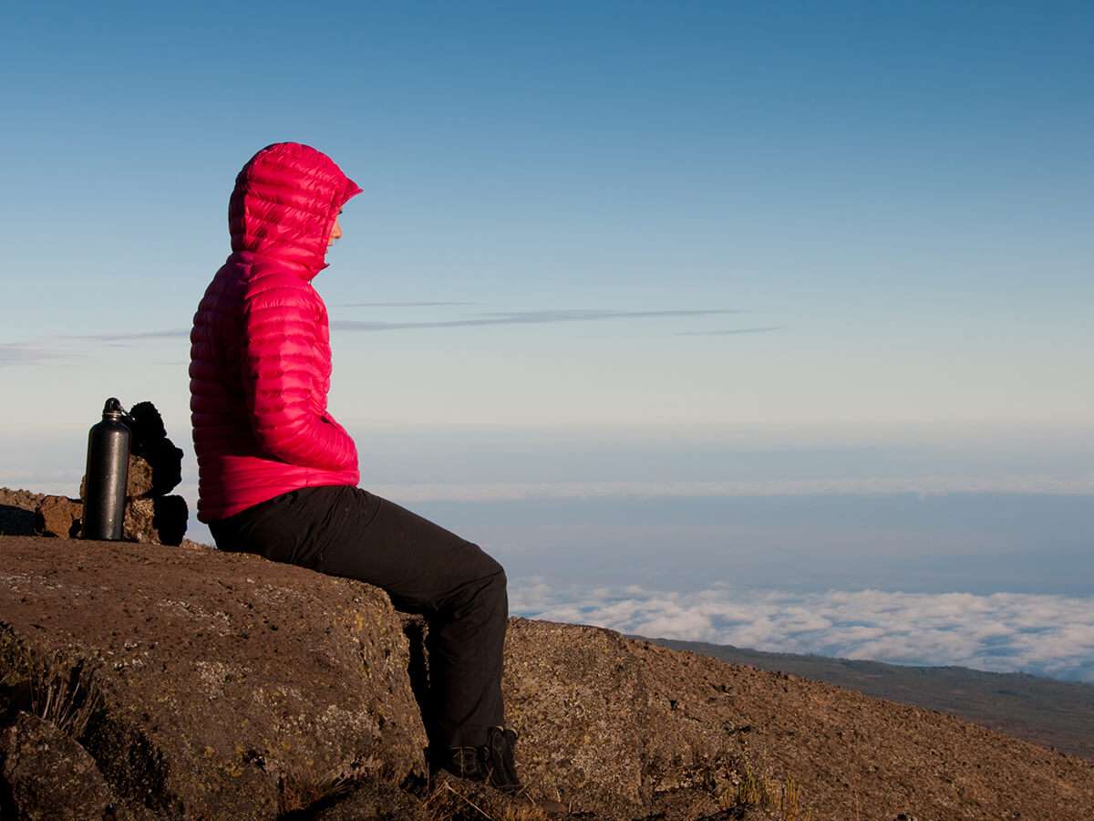 A person in a red jacket sits on a rocky ledge, overlooking the vast, cloud-filled landscape surrounding Mount Kilimanjaro under a clear blue sky. A black thermos is placed nearby on the ground. - Easy Travel Tanzania