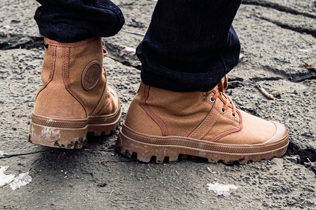 坦桑尼亚 1 号步行靴特写 一个人穿着棕褐色高帮帆布靴和深色裤子,站在一块布满裂纹的灰色地面上。这双靴子是探险旅行必备的物品,如今却沾满污垢和磨损,更显其粗犷的质感。——坦桑尼亚轻松旅行