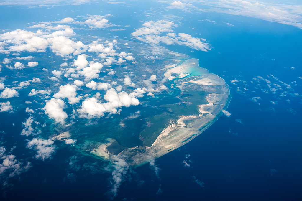 Aerial view of a Tanzanian island surrounded by the ocean, with scattered clouds in the sky. The island features varied landscapes, including sandy beaches and lush greenery. The sea shows different shades of blue, highlighting the coastline. - Easy Travel Tanzania