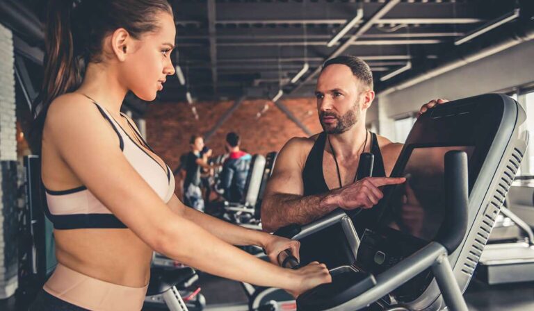 Een vrouw in sportkleding is gefocust op een crosstrainer in de sportschool en belichaamt haar fitnessdoelen. Een man in sportkleding staat naast haar en wijst naar de apparaten. Op de achtergrond zijn andere sportschoolbezoekers te zien die hun routines op soortgelijke apparaten perfectioneren. - Easy Travel Tanzania