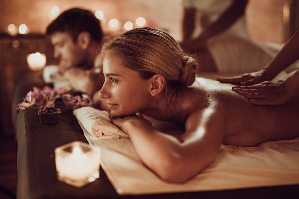 In a dimly lit room reminiscent of Mnemba Islands serenity, two people receive massages on tables, their heads turned sideways. Candles and flowers enhance the tranquil ambiance. The focus is on the person in the foreground, smiling and appearing thoroughly relaxed. - Easy Travel Tanzania