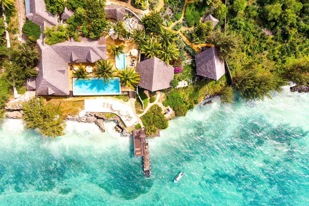 Aerial view of a Mnemba-style tropical resort with thatched-roof buildings and a pool nestled in lush greenery. The resort sits beside a turquoise ocean, with a small pier and boat visible near the shoreline, offering an idyllic escape. - Easy Travel Tanzania