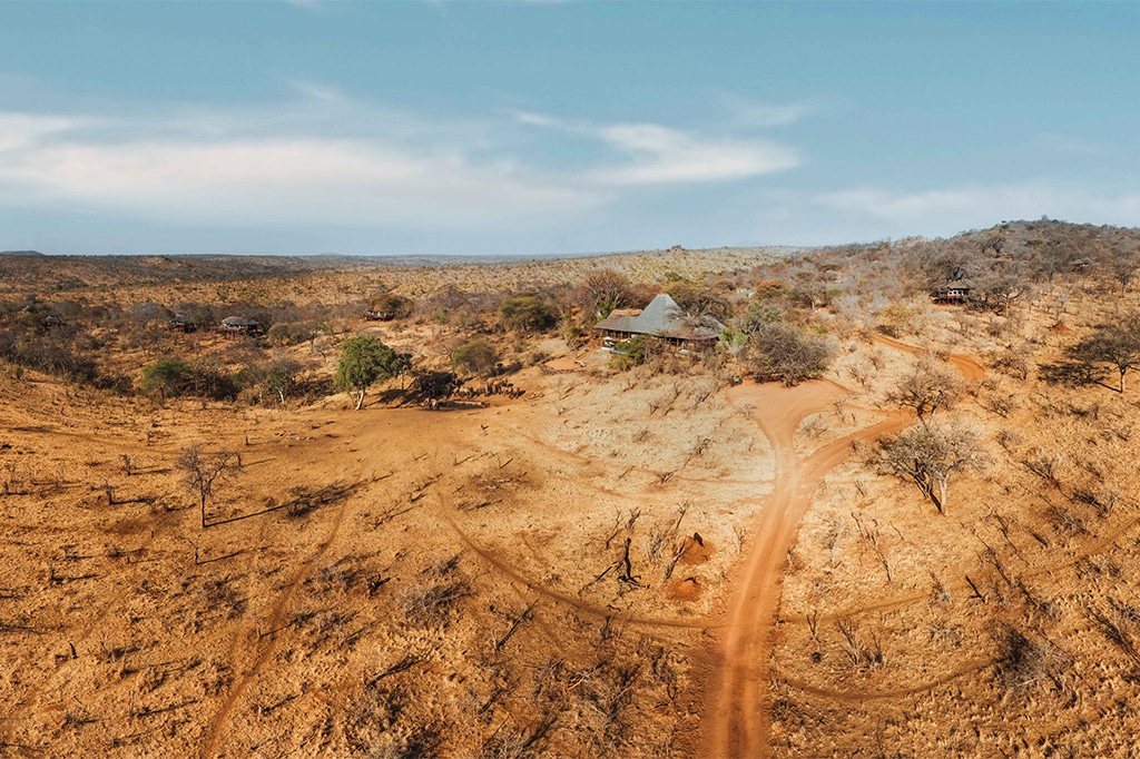 Aerial view of a dry, expansive landscape reminiscent of tarangire national park, with sparse vegetation and a dirt road leading to a few small structures nestled among trees. The sky is mostly clear, with a few clouds casting light shadows on the terrain. - easy travel tanzania