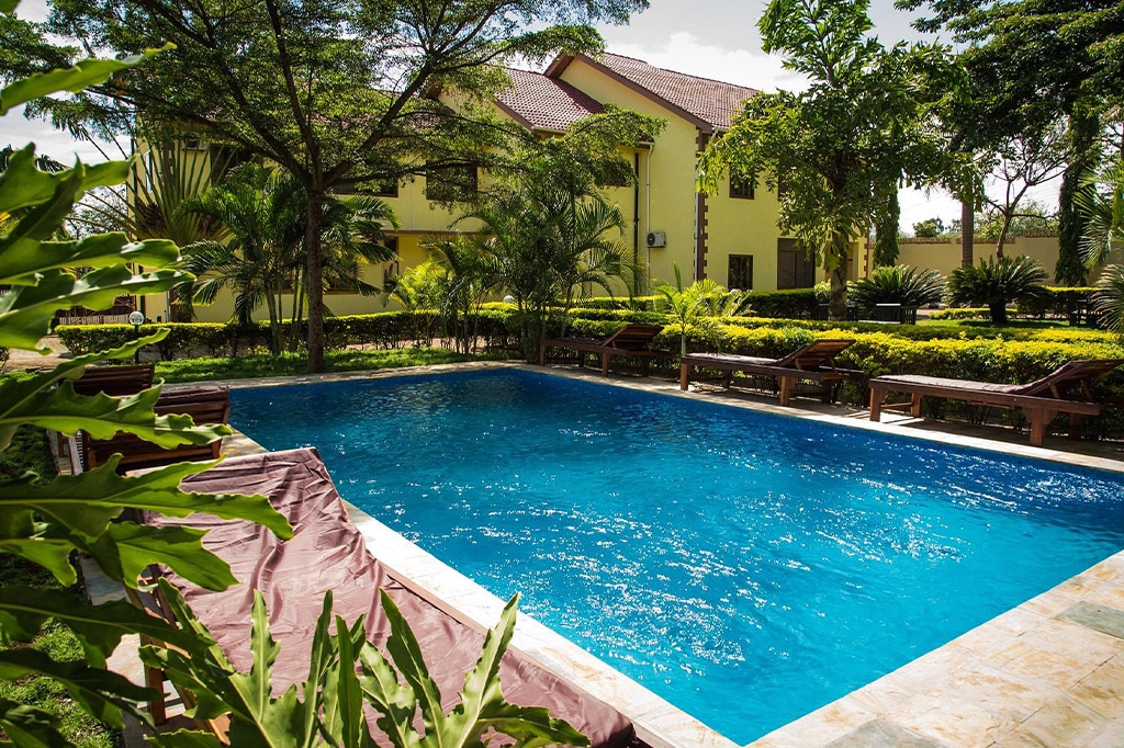 A serene outdoor swimming pool, surrounded by lush greenery and sunbeds under large trees, exudes a tranquil moshi ambiance. A yellow building peeks from behind the foliage, partially shaded in this sunny, peaceful oasis. - Easy Travel Tanzania