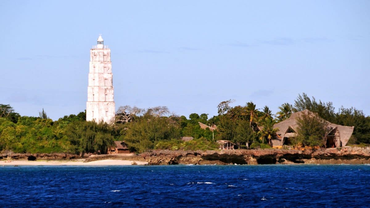 Un faro bianco si erge su una costa rocciosa, circondato da alberi verdi e poche capanne con il tetto di paglia. Il mare azzurro si estende in primo piano, sotto un cielo terso. - Easy Travel Tanzania