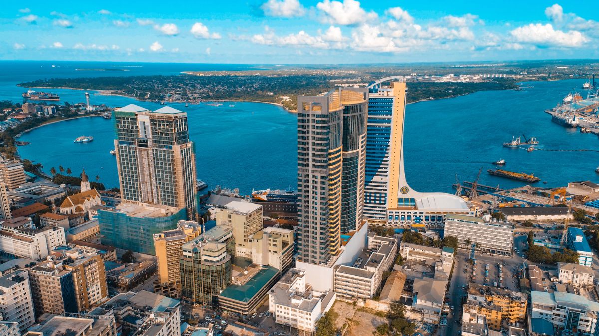 Aerial view of a city skyline featuring modern high-rise buildings along a waterfront. The blue sea is visible in the background with boats and a distant coastline under a clear sky, dotted with clouds. Urban streets and smaller structures are in the foreground. - Easy Travel Tanzania