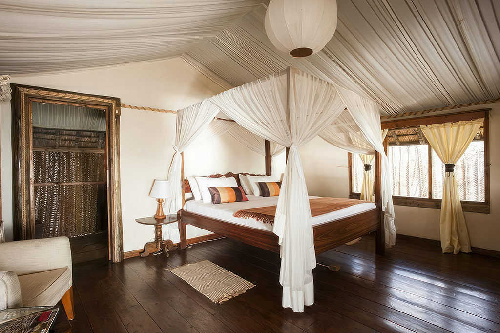 In de kamer van Saadani Safari Lodge Tanzania 1 Luxe slaapkamer die doet denken aan een verblijf in het Saadani National Park, met een hemelbed gedrapeerd in transparante witte stof met een houten frame. De kamer heeft een gepolijste houten vloer, een tapijt, warme verlichting, nachtkastjes met lampen en een groot raam met gordijnen. - Easy Travel Tanzania