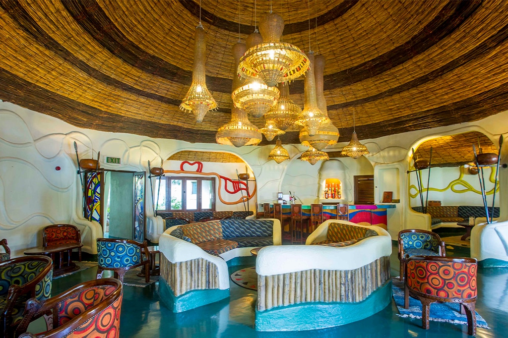 Een levendige en artistieke lounge, geïnspireerd door Lake Manyara, met kleurrijke stoelen en gebogen houten banken in een cirkelvormig patroon. Hangende, mandachtige lampen dalen neer aan het rieten plafond, terwijl de muren zijn voorzien van abstracte ontwerpen en een mix van patronen. - Easy Travel Tanzania