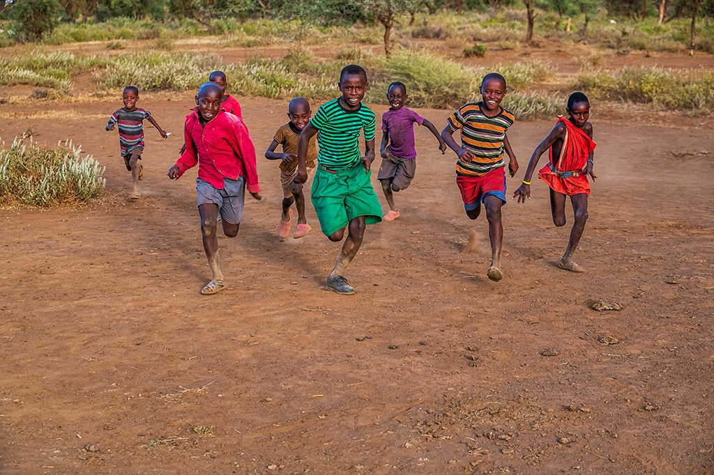 A group of children joyfully running on a dirt path in an outdoor setting at saadani national park, wearing colorful clothing. The background features trees and shrubs, suggesting a rural environment. The children appear to be smiling and engaged in playful activity. - easy travel tanzania