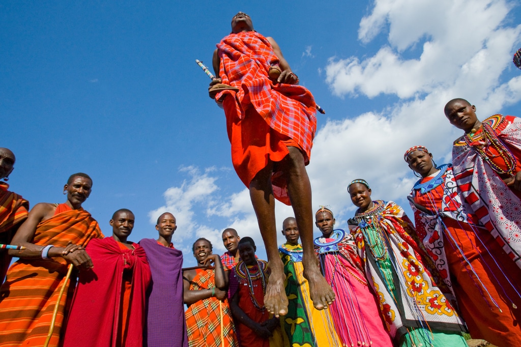 Un uomo Masai in abiti tradizionali salta in aria durante una cerimonia tradizionale vicino a Ndutu, circondato da uomini e donne Masai in abiti colorati e gioielli. Il cielo azzurro e le nuvole fanno da sfondo a questa vibrante celebrazione culturale. - Easy Travel Tanzania