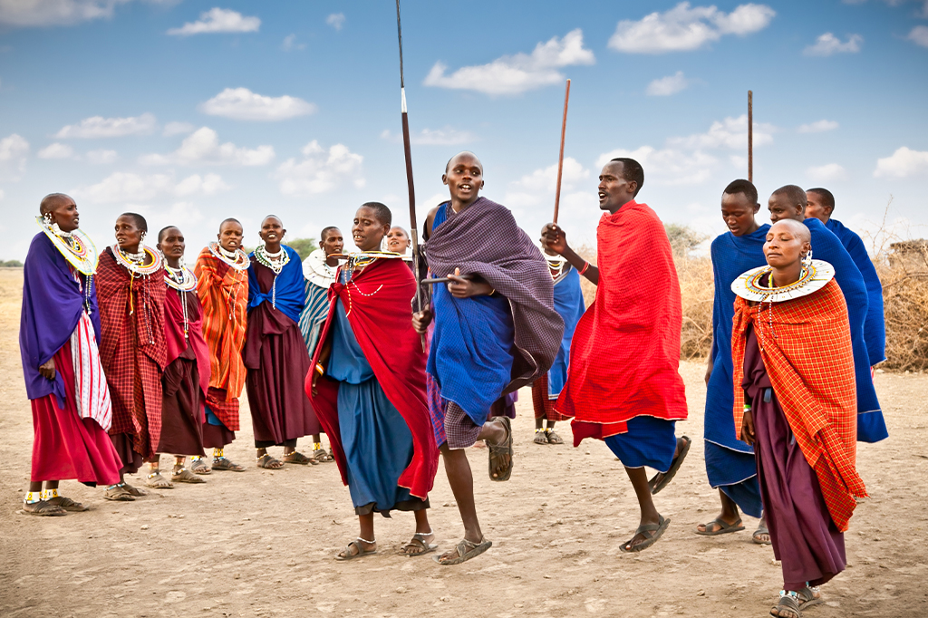 A group of people dressed in colorful traditional attire gather outdoors in the vibrant serengeti national park. Some jump joyfully, while others watch and hold wooden staffs. The clear sky with scattered clouds adds to the lively scene. - easy travel tanzania