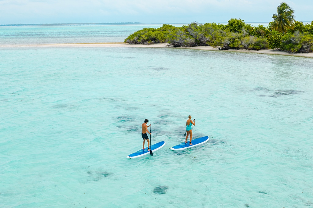 Deux personnes font du paddle sur une eau turquoise et limpide près de l'île de Mnemba. Le ciel est nuageux et une végétation luxuriante est visible sur l'île en arrière-plan. - Easy Travel Tanzania