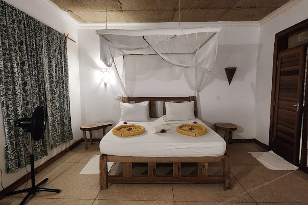 В уютной спальне в Кизимкази Димбани стоит кровать с деревянным каркасом, белое постельное белье и лёгкий балдахин. На двух круглых тканых ковриках украшены свёрнутые полотенца и декоративные цветы. Слева – узорчатая занавеска и напольный вентилятор, а тёплое освещение создаёт уютную атмосферу. - Easy Travel Tanzania