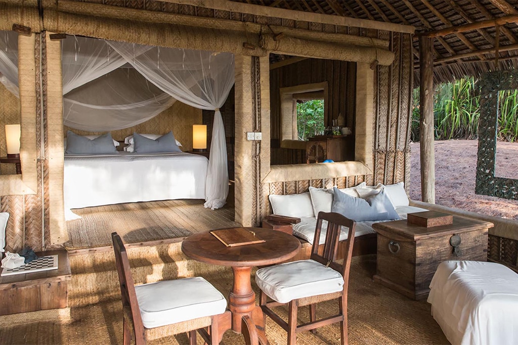 Una acogedora habitación tropical en la isla de Mnemba cuenta con una cama con dosel cubierta con una mosquitera blanca. La habitación incluye muebles de madera natural, una pequeña mesa de comedor con sillas, iluminación tenue y una vista al aire libre rodeada de exuberante vegetación. - Easy Travel Tanzania