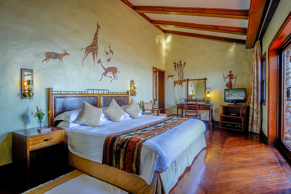 Luxe hotelkamer met een groot bed, versierd met kleurrijke kussens en een deken. De kamer is voorzien van muurschilderingen van giraffen en antilopen, die doen denken aan de rijke fauna van de Ngorongoro. Een houten vloer, tv en bureau maken de ruimte compleet. Grote ramen laten natuurlijk licht binnen en benadrukken de serene inrichting. - Easy Travel Tanzania