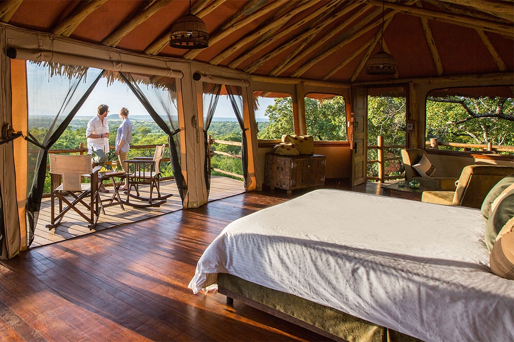 A luxurious, open-air hotel room with a large bed and wooden floors, overlooking a scenic view in tarangire national park. Two people stand outside on the deck, surrounded by lush greenery and distant hills under the clear sky. - easy travel tanzania