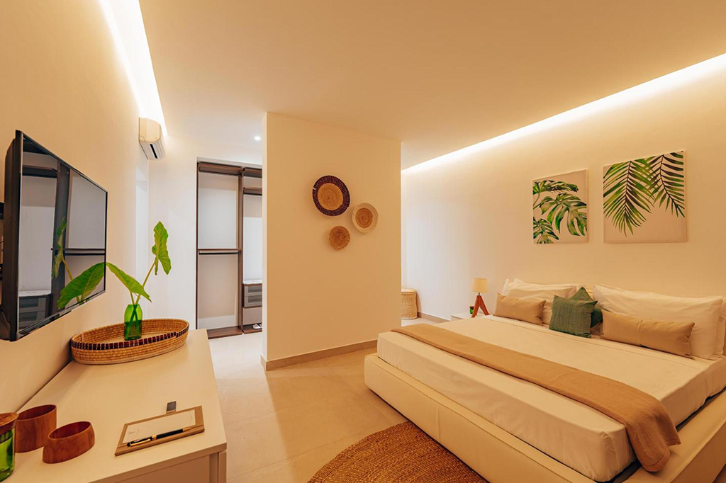 Un acogedor dormitorio con un diseño moderno inspirado en Mnemba cuenta con una cama grande con sábanas blancas y beige. La decoración incluye dos láminas con motivos de hojas, cestas de pared y un televisor. La suave iluminación realza la cálida atmósfera, complementada por una maceta sobre la cómoda. - Easy Travel Tanzania