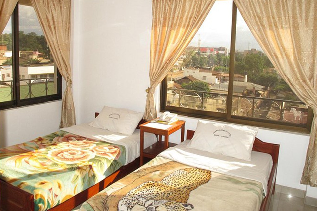 A cozy hotel room with two single beds, each adorned with colorful blankets—one floral and the other featuring a leopard design. Large windows with golden curtains offer a view of Moshis picturesque buildings and greenery outside. A small nightstand sits between the beds. - Easy Travel Tanzania