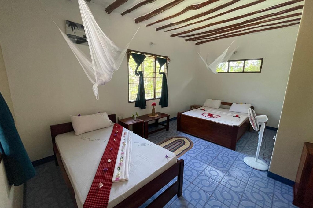 Уютная комната с двумя аккуратно застеленными кроватями с белым постельным бельем, украшенная красными акцентами, воссоздает очарование Кизимкази. Деревянный потолок и кафельный пол располагают к спокойствию, а окна с зелёными шторами и москитные сетки создают уютную атмосферу. Напольный вентилятор охлаждает помещение, а плетёный коврик дополняет интерьер. - Easy Travel Tanzania