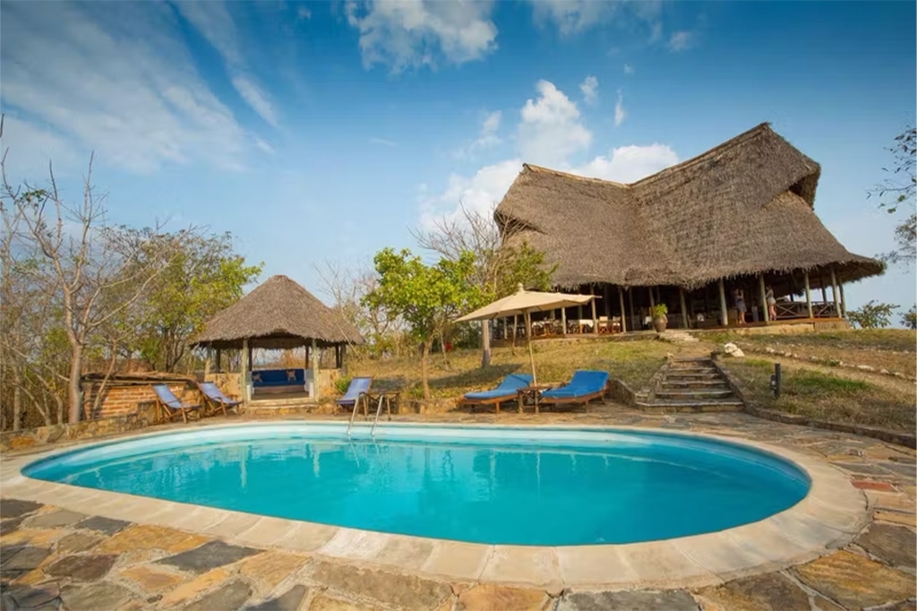 石造りのパティオに囲まれた静かな屋外プールには、青いラウンジチェアとパラソルがいくつか置かれている。背景には、ミクミ国立公園を思わせる素朴な茅葺き屋根のロッジが、雲が散らばる青空の下、草に覆われた丘の上に佇んでいる。 - Easy Travel Tanzania