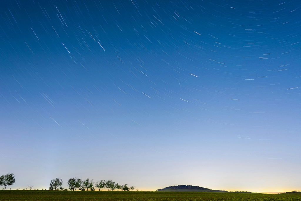 A long-exposure photo captures star trails forming circular patterns over the serengeti national park, with a row of trees and a distant hill silhouetted against the gentle gradient of the night skys lower half, blending from deep blue to light. - easy travel tanzania