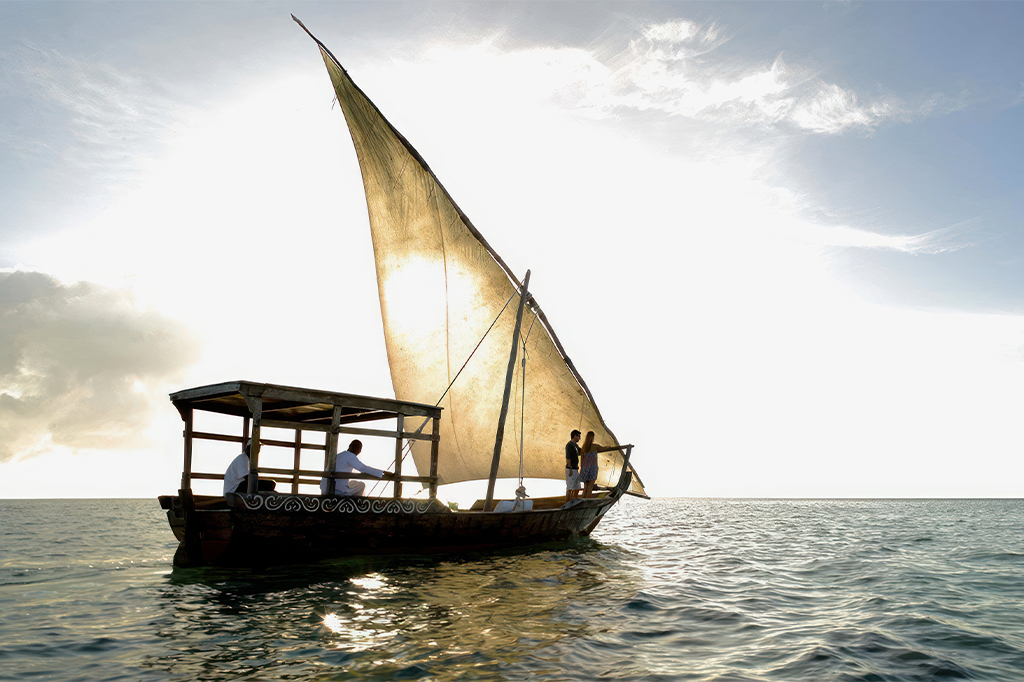 一艘传统的单桅帆船，挂着一面巨大的单帆，在慵懒泻湖岛附近阳光明媚的海面上缓缓驶过。船上载着几个人，在明亮的天空映衬下，映衬出他们的轮廓。平静的水面倒映着柔和的阳光，营造出一幅宁静祥和的景象。——坦桑尼亚轻松旅行