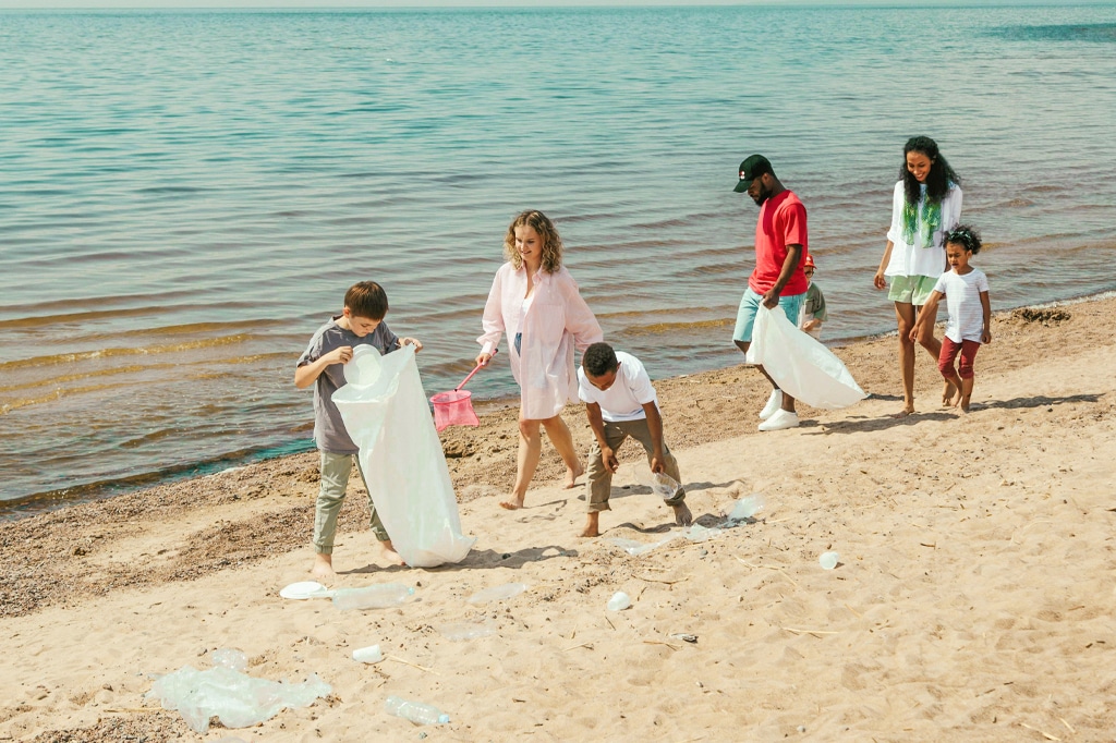 A group of people, including children and adults, are collecting litter on Dimbani Mkunguni Beach near calm water. They are using large plastic bags for the cleanup. The sky is clear, and the atmosphere is cheerful and collaborative. - Easy Travel Tanzania