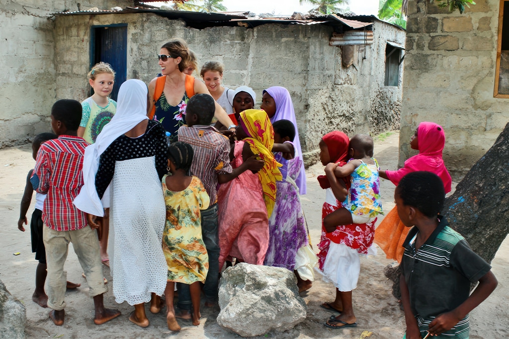 一群孩子和两个成年人一起站在户外的石头建筑前，让人想起风景如画的乌宗瓦山脉。孩子们穿着色彩鲜艳的裙子和帽子。一位成年人戴着墨镜，图片右侧可见一棵树干。——坦桑尼亚轻松旅行