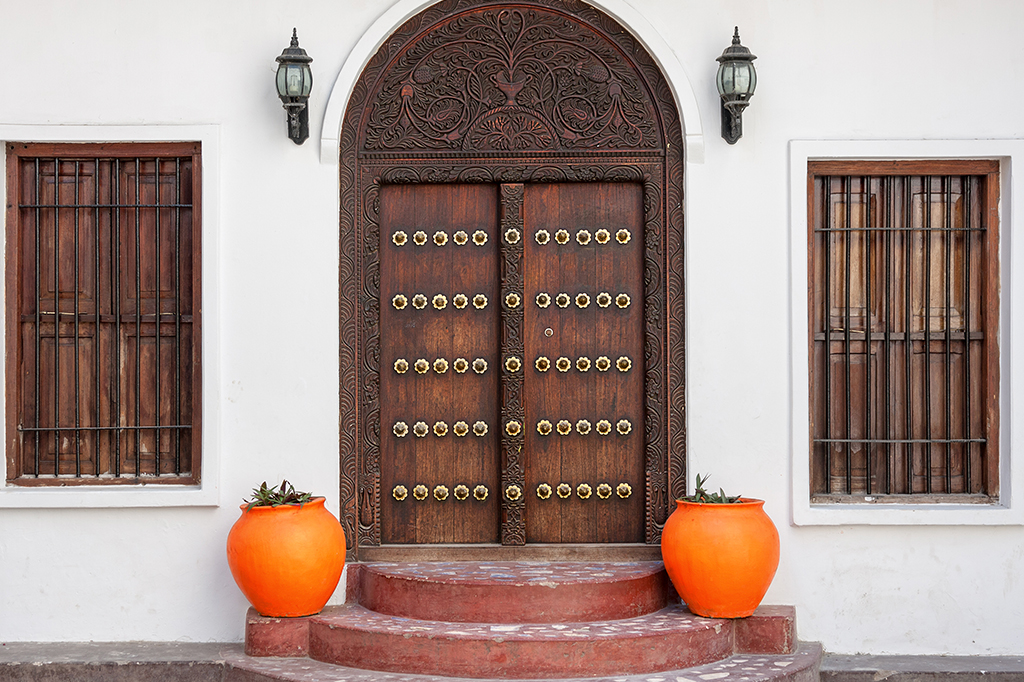 Eine hölzerne Doppeltür mit kunstvollen Schnitzereien und Messingbeschlägen erinnert an den Charme von Stone Town, eingerahmt von zwei Bogenfenstern mit Holzgittern. Zwei große, leuchtend orangefarbene Töpfe flankieren den Eingang, über der Tür sind zwei Wandlampen angebracht. – easy travel tanzania