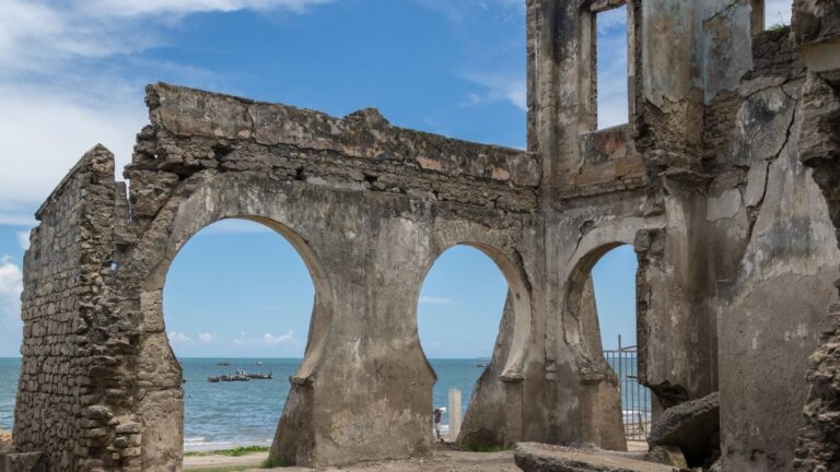 Ein verwittertes, bröckelndes Steingebäude mit drei großen Bögen überblickt ein ruhiges Meer und entfernte Boote unter einem blauen Himmel mit vereinzelten Wolken. - Easy Travel Tanzania