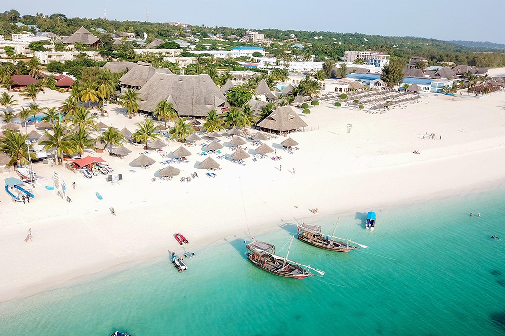 Aerial view of Kendwas tropical beach with white sand and turquoise water. Traditional wooden boats are anchored near the shore, while thatched-roof buildings and beach umbrellas with loungers nestle under palm trees, framed by lush greenery. - Easy Travel Tanzania