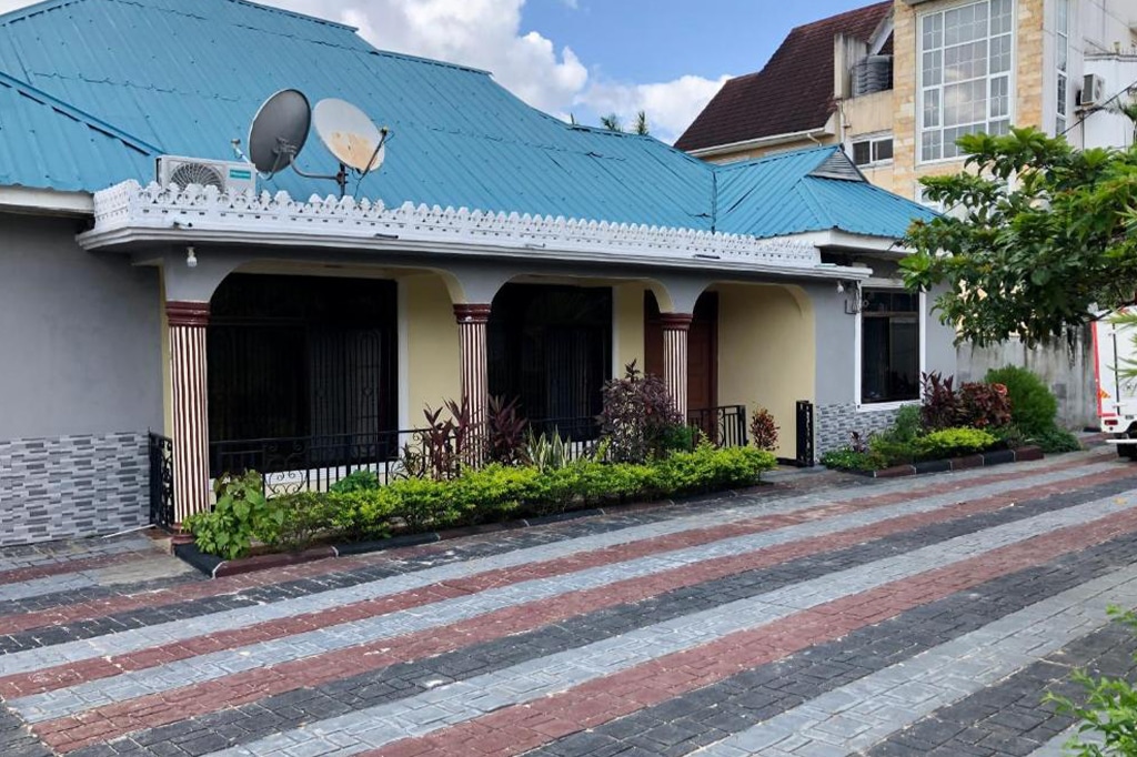 A single-story house in dar es salaam boasts a blue metal roof with two satellite dishes. The facade features arches, a mix of paint colors, and brick-like tiles. Small shrubs line the front, bordered by a paved pathway adorned with red, black, and gray tiles. - easy travel tanzania