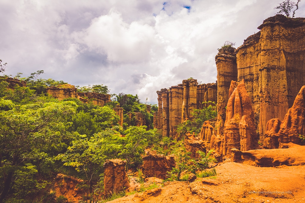 A scenic view of red rock canyons with lush green vegetation reminiscent of Iringas landscapes. The towering formations stand under a partly cloudy sky, creating a contrast between the earth-toned cliffs and vibrant greenery. - Easy Travel Tanzania