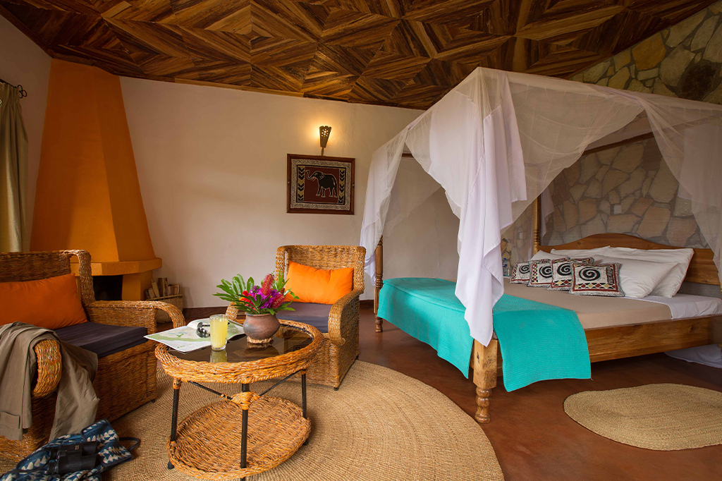 Room Country Lodge Karatu Tanzania 1 A cozy room in Karatu featuring a canopy bed with white netting and vibrant turquoise covers, wicker furniture with orange cushions, and a round wicker table adorned with flowers. The walls showcase art, enhancing the warm ambiance from the earthy decor. - Easy Travel Tanzania