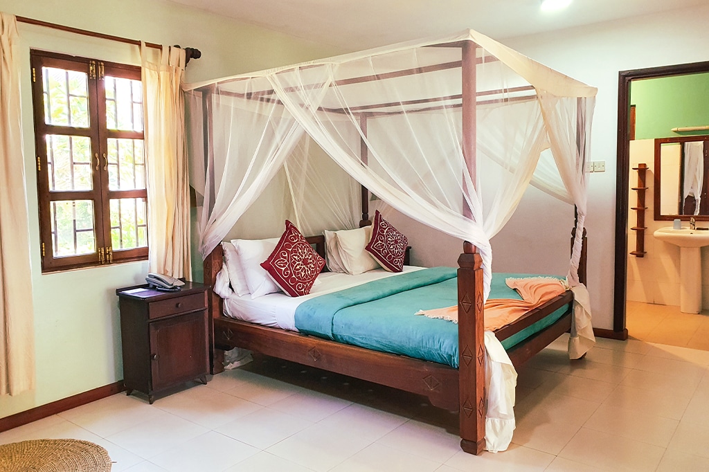 A cozy Kendwa-inspired bedroom features a wooden four-poster bed with white mosquito netting and teal bedding. Two red pillows with intricate designs adorn the bed. The room has soft lighting, a window with curtains, and a glimpse of an adjacent bathroom. - Easy Travel Tanzania
