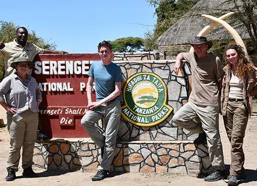 Groupe de personnes posant devant le panneau d'entrée du parc national du Serengeti, en Tanzanie, avec l'emblème du parc et un fond rustique.