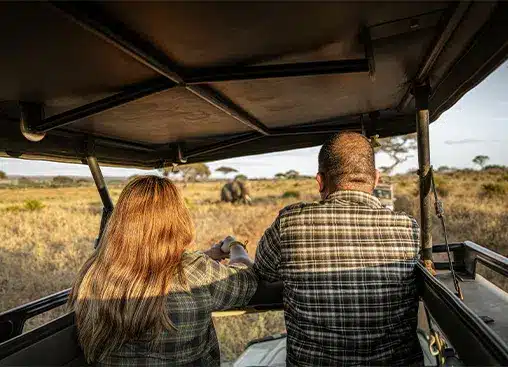 Twee mensen in een safarivoertuig observeren een olifant in een open savannelandschap onder een heldere hemel.