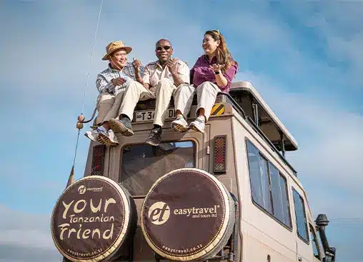 Three people sit atop a safari vehicle with "Your Tanzanian Friend" and "easy travel" logos, enjoying a scenic view.