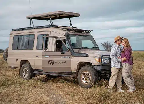 Una pareja se abraza junto a un vehículo de safari en un paisaje herboso, bajo un cielo nublado.