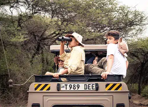 Safari tourists in a jeep, observing wildlife with binoculars, surrounded by lush greenery and trees.