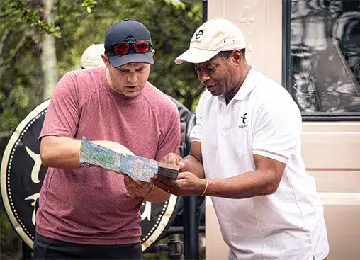 Due uomini all'aperto esaminano insieme una mappa, uno con una camicia rossa e un berretto, l'altro con una polo bianca e un cappello, vicino a un veicolo.