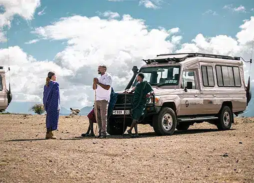 Guía de safari y turistas cerca de un vehículo 4x4 en un vasto paisaje abierto bajo un cielo azul con nubes.